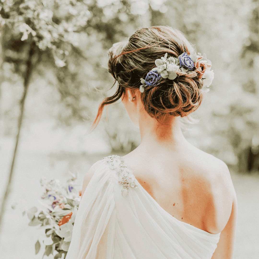 Bride with floral hairpiece and draped gown, standing outdoors amidst greenery.