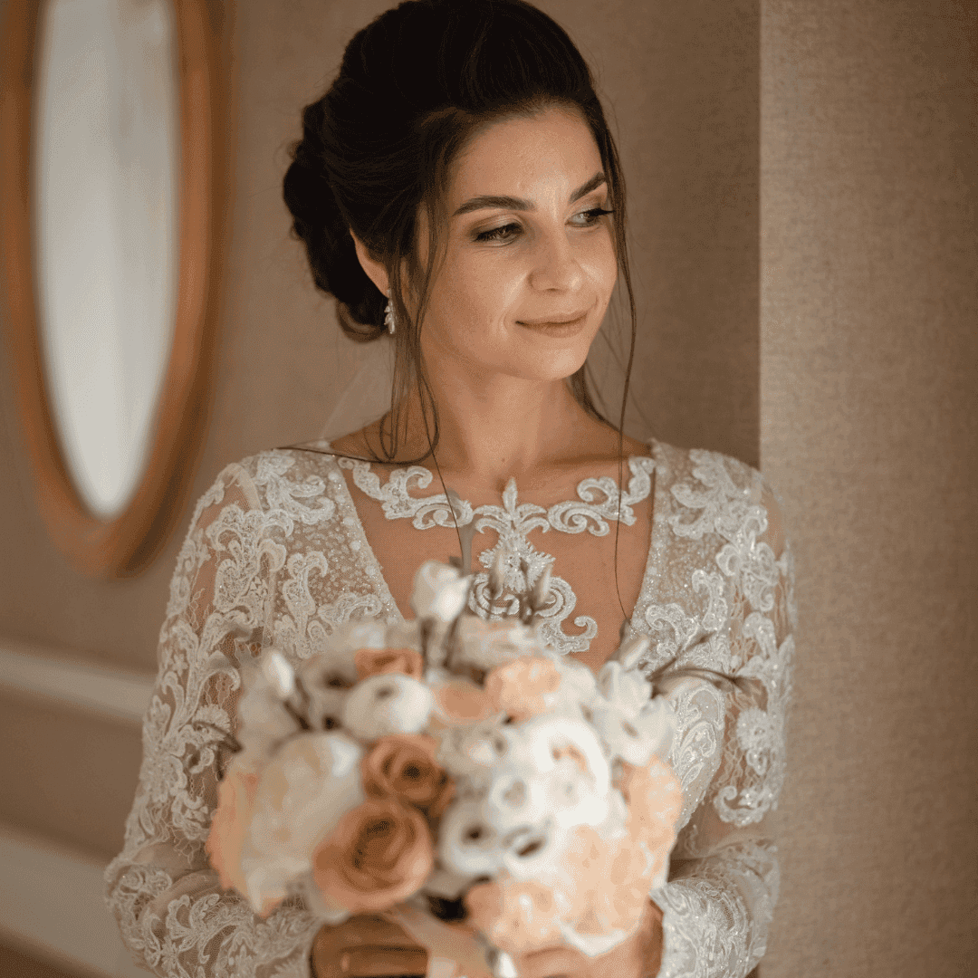 Bride in lace wedding dress holding a bouquet of white and peach roses, smiling softly.